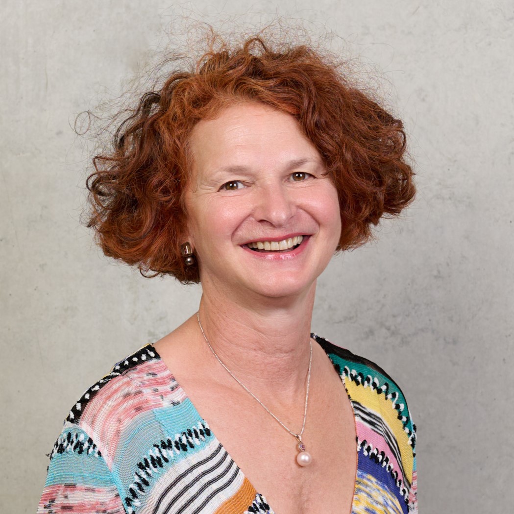 Eine lächelnde Frau mit roten Locken, die ein bunt gestreiftes Oberteil und eine silberne Kette mit einem Perlenanhänger trägt, posiert vor einem hellgrauen, strukturierten Hintergrund - Antje Vondran strahlt Selbstbewusstsein und Charme aus. at Hochschule Coburg