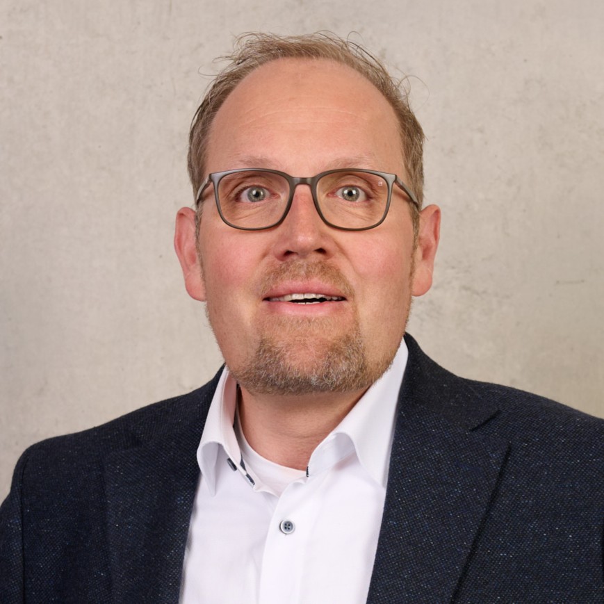 Ein Mann mit kurzen blonden Haaren, Brille und einem gepflegten Bart trägt einen dunklen Blazer über einem weißen Hemd und posiert vor einem hellen, schlichten Hintergrund – das ist Matthias J. Kaiser. at Hochschule Coburg