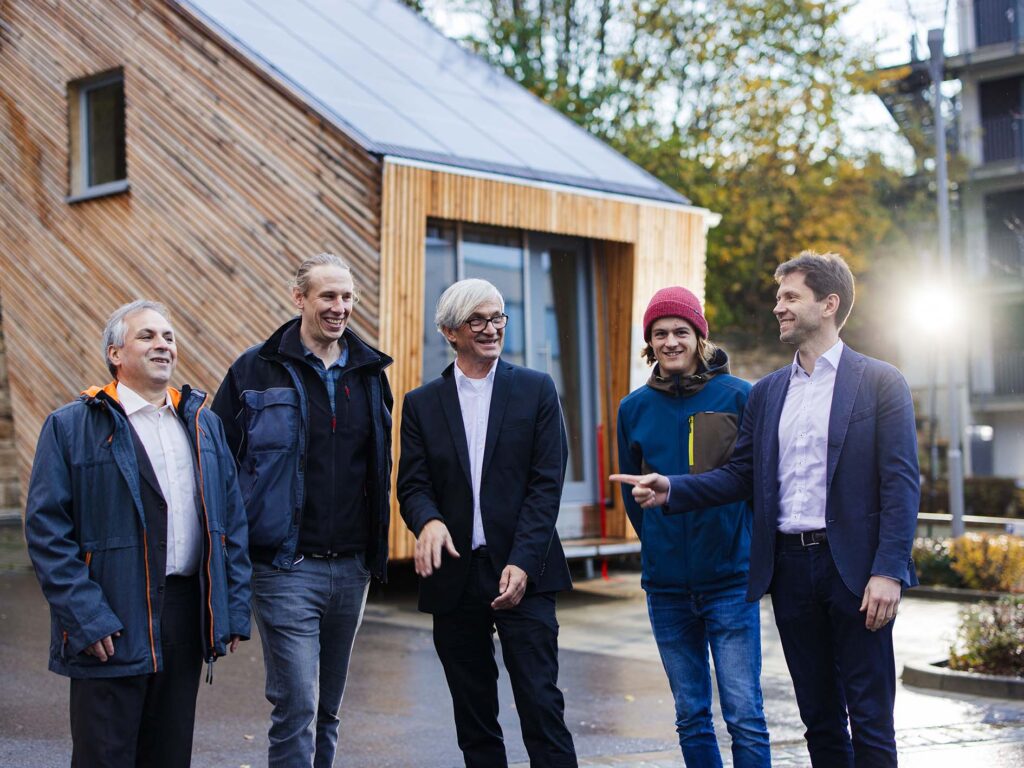 Fünf Menschen stehen vor einem modernen Holzgebäude in der Nähe der Hochschule Coburg, lachen und unterhalten sich. Sie sind leger gekleidet und tragen Jacken, einer trägt eine rote Mütze. Im Hintergrund ist eine sonnige, grüne Straßenszene zu sehen, die zur lebhaften Atmosphäre beiträgt.