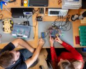 Zwei Personen arbeiten an einem Schreibtisch voller elektronischer Werkzeuge und Komponenten und konzentrieren sich auf eine Leiterplatte. Daneben stehen ein Roboterchassis auf Rädern, ein Multimeter und ein Oszilloskop. Das Gemeinschaftsprojekt wirkt wie ein praxisorientierter Workshop, der typisch für den innovativen Lernansatz der Hochschule Coburg ist.