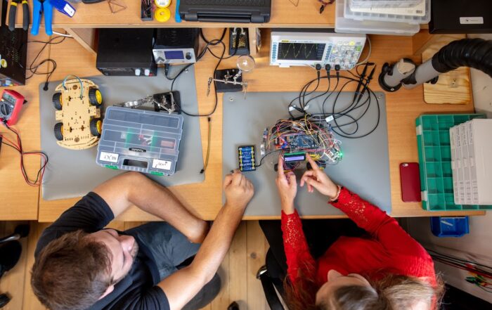 Zwei Personen arbeiten an einem Schreibtisch voller elektronischer Werkzeuge und Komponenten und konzentrieren sich auf eine Leiterplatte. Daneben stehen ein Roboterchassis auf Rädern, ein Multimeter und ein Oszilloskop. Das Gemeinschaftsprojekt wirkt wie ein praxisorientierter Workshop, der typisch für den innovativen Lernansatz der Hochschule Coburg ist.