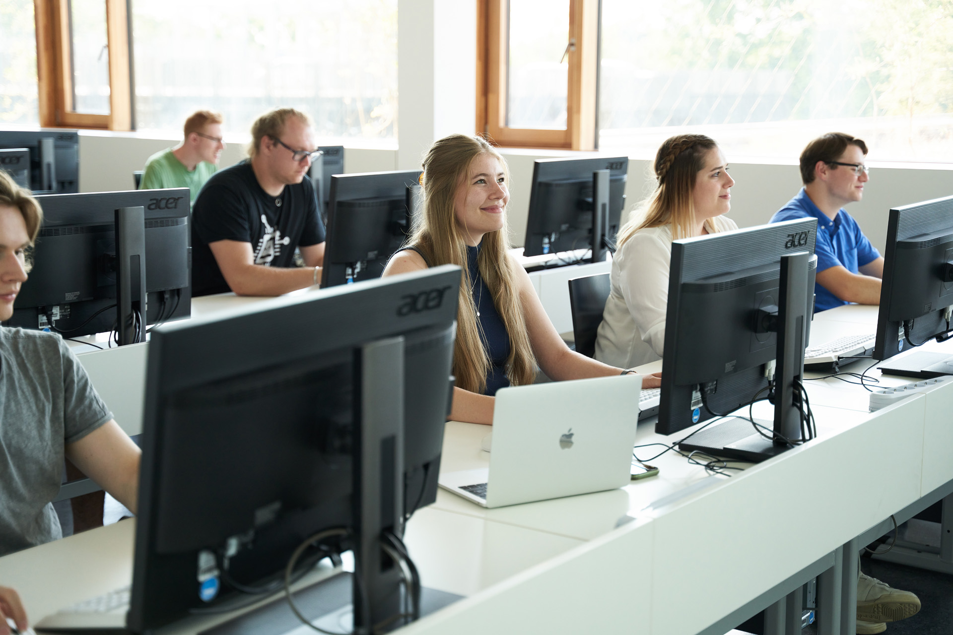 In einem Hörsaal der Hochschule Coburg sitzen Studierende an Tischreihen und arbeiten am Computer. Sie wirken konzentriert und aufmerksam. Große Fenster im Hintergrund lassen Tageslicht herein und schaffen eine helle und luftige Atmosphäre.