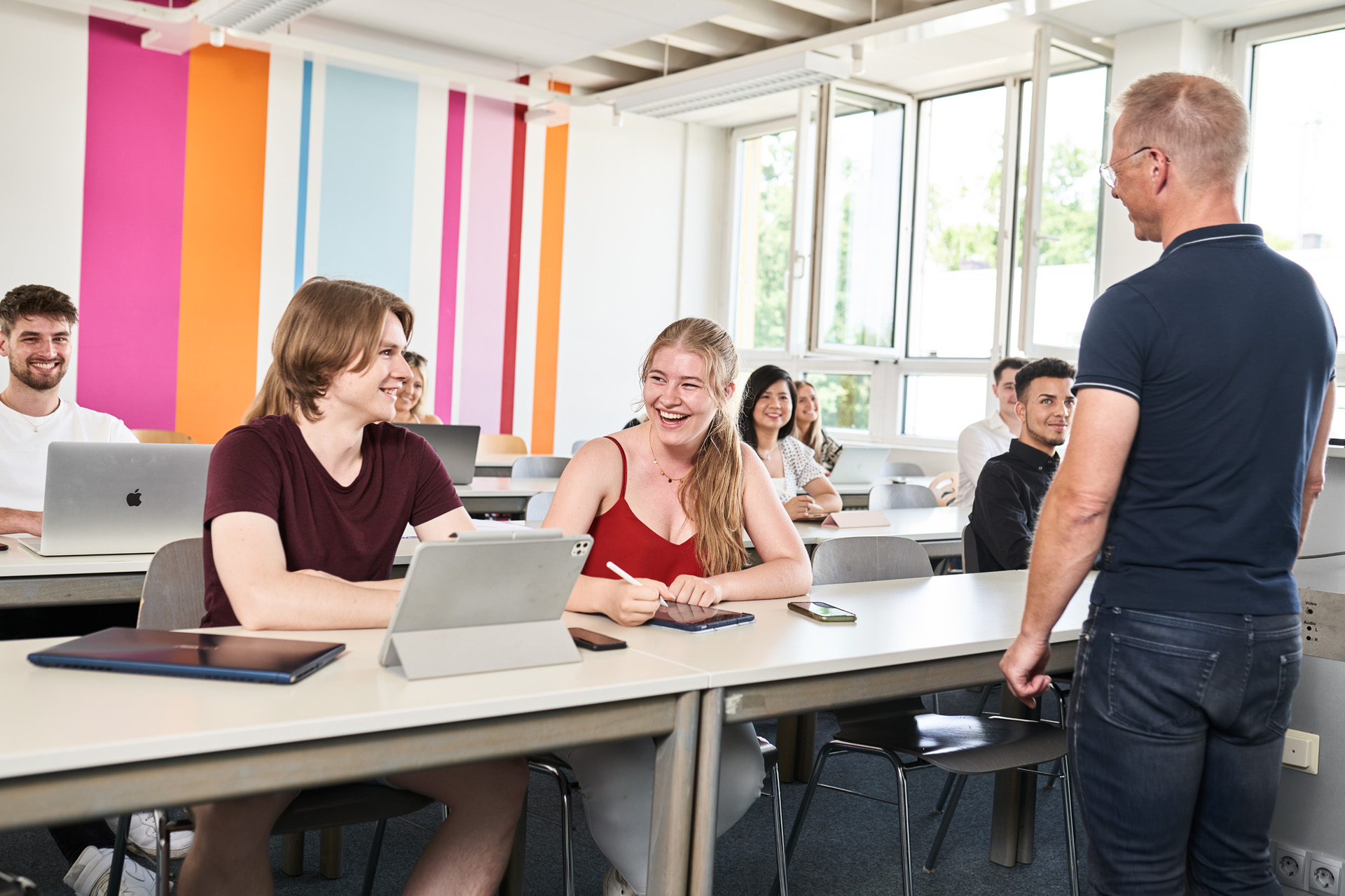 Mehrere Studierende in einem Seminarraum, die sich gegenseitig anlachen, im Vordergrund der Dozent der sie anlächelt