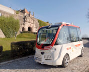 Ein kleines, fahrerloses Shuttle mit rot-weißem Design steht auf einem Kopfsteinpflasterweg in der Nähe eines alten Steingebäudes der Hochschule Coburg. Der Himmel ist klar und Bäume mit spärlichem Laub säumen die Straße und fangen die Harmonie von Tradition und Innovation ein.