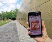 Eine Hand hält ein Smartphone mit einer E-Mail zum Thema „25 Jahre“ vor einem modernen, perforierten Metallgebäude der Hochschule Coburg. Die Szene ist im Freien, mit Bäumen und einem bewölkten Himmel im Hintergrund.