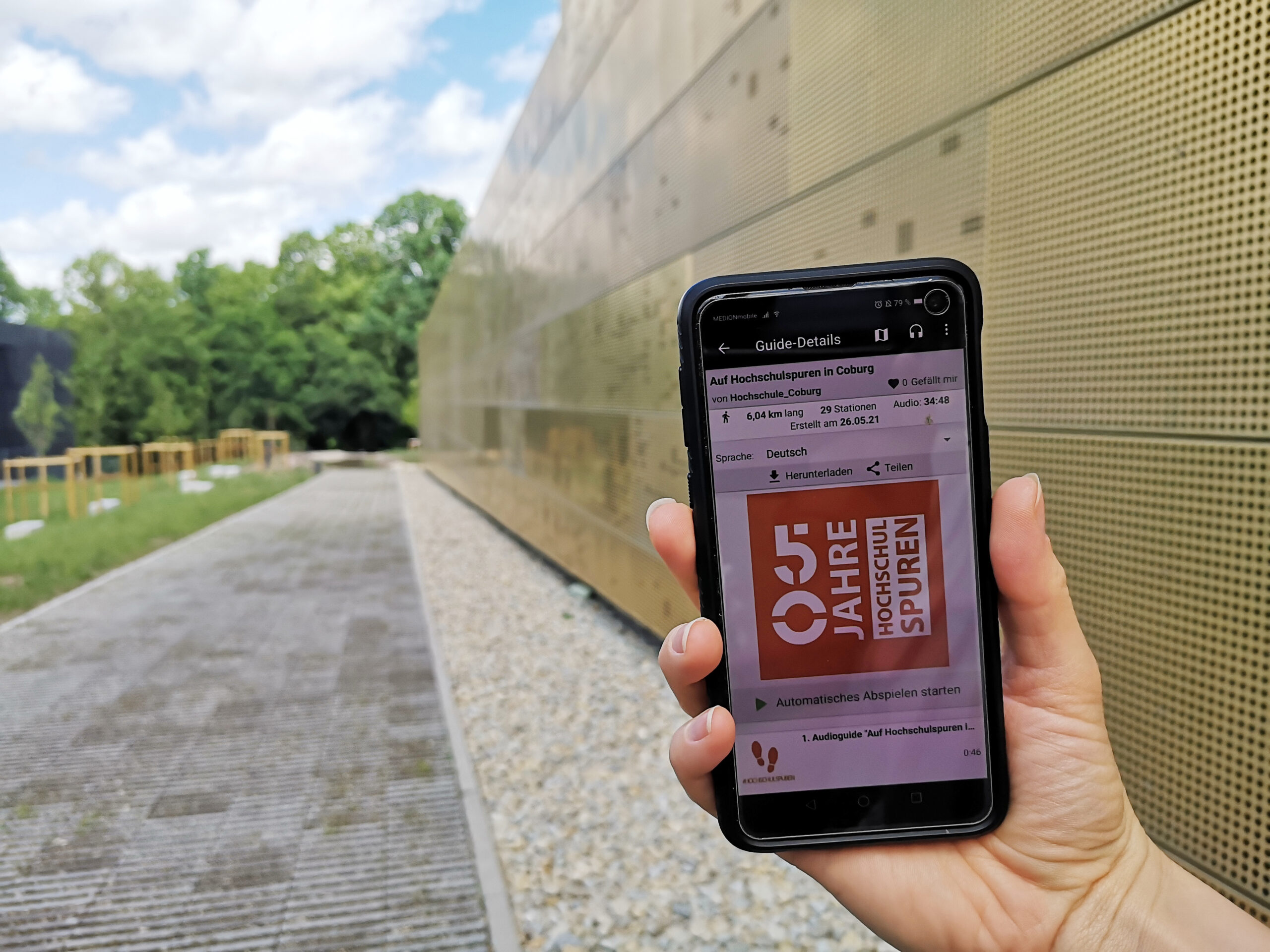 Eine Hand hält ein Smartphone mit einer E-Mail zum Thema „25 Jahre“ vor einem modernen, perforierten Metallgebäude der Hochschule Coburg. Die Szene ist im Freien, mit Bäumen und einem bewölkten Himmel im Hintergrund.