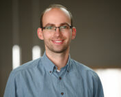 Ein Mann mit Brille und Bart lächelt und trägt ein blaues Hemd mit Knöpfen. Der sanft beleuchtete Hintergrund mit vertikalen Lichtlinien lässt vermuten, dass er an der Hochschule Coburg ist und sich vielleicht auf eine spannende Vorlesung oder eine Campus-Veranstaltung vorbereitet.