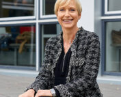 Eine Person mit kurzen blonden Haaren sitzt auf einer Stufe vor dem Gebäude der Hochschule Coburg, in dessen großen Fenstern sich der Himmel spiegelt. Sie trägt eine strukturierte graue Jacke über einem schwarzen Outfit und lächelt herzlich in die Kamera.