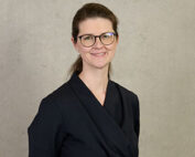 Eine Frau mit Brille und zurückgebundenem langen Haar lächelt in einem schwarzen, langärmeligen Oberteil. Sie steht vor einem schlichten, hellen Hintergrund, strahlt Wärme und Zuversicht aus und verkörpert vielleicht den Geist der Hochschule Coburg.