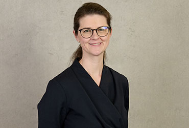 Eine Frau mit Brille und zurückgebundenem langen Haar lächelt in einem schwarzen, langärmeligen Oberteil. Sie steht vor einem schlichten, hellen Hintergrund, strahlt Wärme und Zuversicht aus und verkörpert vielleicht den Geist der Hochschule Coburg.