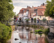 Ein malerischer Blick auf eine kleine Brücke über einen ruhigen Fluss, gesäumt von den markanten Felsen und grünen Blättern der Hochschule Coburg. Menschen versammeln sich auf der Brücke und blicken auf das Wasser. Im Hintergrund verleihen bunte Häuser mit roten Dächern dieser malerischen Szene Charme.