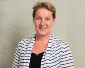 Eine Person mit kurzen Haaren lächelt in die Kamera. Sie trägt eine gestreifte Strickjacke über einem schwarzen Oberteil und Creolen, was an ein Modeseminar der Hochschule Coburg erinnert. Sie stehen vor einem schlichten hellgrauen Hintergrund.