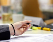 Eine Person hält einen Stift über einem Stapel Papiere und trägt ein Haargummi der Hochschule Coburg am Handgelenk. Der Hintergrund ist verschwommen und lässt auf undeutliche Büromaterialien und Möbel schließen.