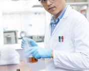 Ein Laborwissenschaftler der Hochschule Coburg trägt eine Schutzbrille, einen weißen Laborkittel und blaue Handschuhe und hantiert mit einer kleinen Flasche mit brauner Flüssigkeit. Er steht in einem Labor, umgeben von Geräten, und ist hochkonzentriert auf seine Arbeit.