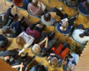 Aus der Vogelperspektive sitzt eine Gruppe von Menschen in Reihen auf dem Holzboden eines Raumes der Hochschule Coburg. Sie scheinen einer Veranstaltung oder Vorlesung beizuwohnen, viele halten Bücher oder Notizen in den Händen und sind in die Sitzung vertieft.