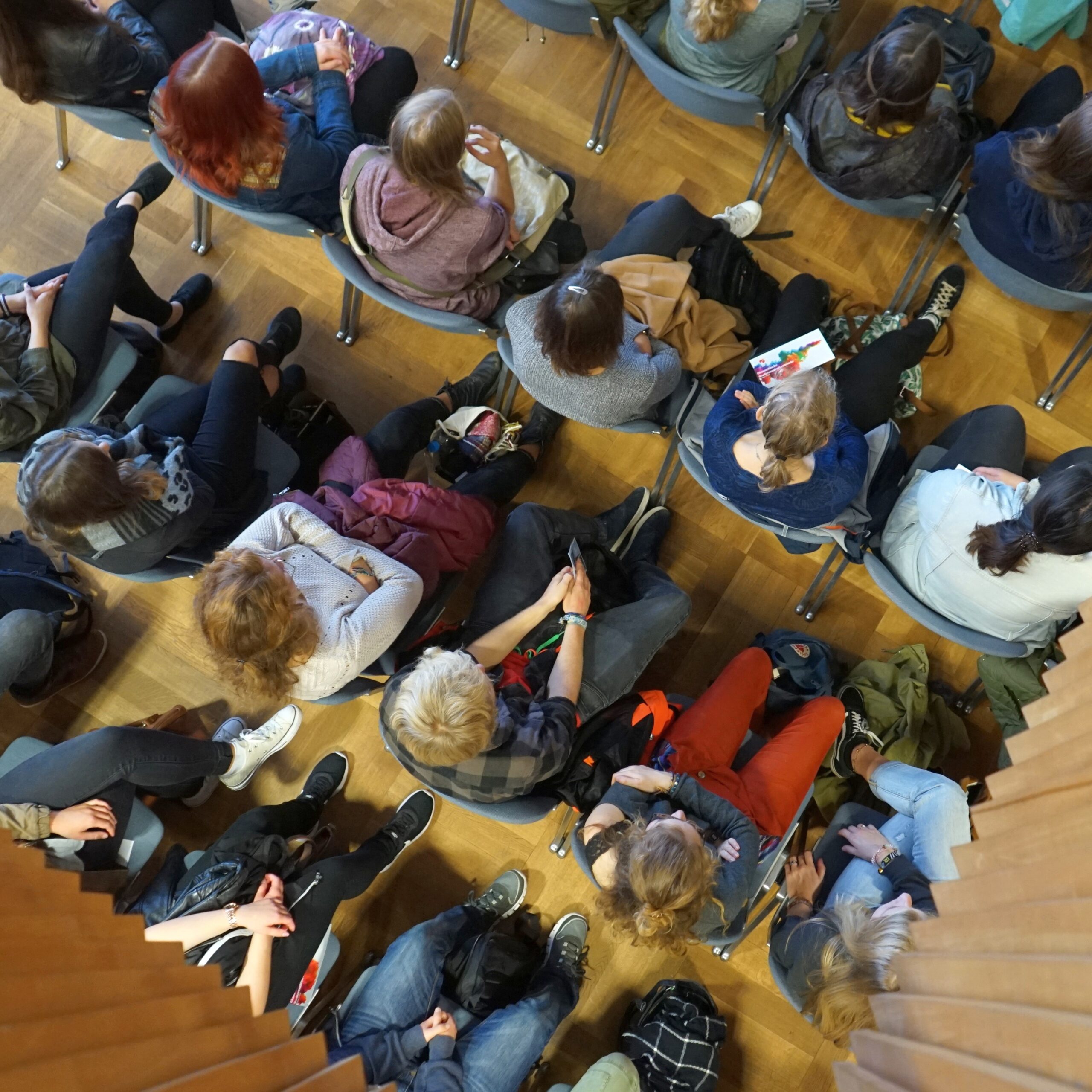 Aus der Vogelperspektive sitzt eine Gruppe von Menschen in Reihen auf dem Holzboden eines Raumes der Hochschule Coburg. Sie scheinen einer Veranstaltung oder Vorlesung beizuwohnen, viele halten Bücher oder Notizen in den Händen und sind in die Sitzung vertieft.