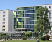 Ein modernes Wohngebäude mit grün-weißen Fassadenpaneelen in der Nähe der Hochschule Coburg ist von Bäumen umgeben. Im Vordergrund stehen Fahrräder unter einer Überdachung, während Menschen auf der Wiese unter einem klaren blauen Himmel ein Sonnenbad nehmen.