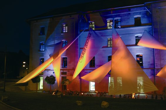 Ein Gebäude, das nachts von bunten Lichtern angestrahlt wird und mit dreieckigen Stoffstrukturen geschmückt ist, auf denen „concrete dreams“ und der Name Hochschule Coburg zu sehen sind. Die Darstellung erzeugt eine lebendige und künstlerische Atmosphäre.
