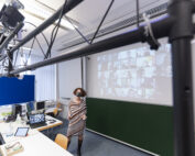 Eine Person steht in einem Hörsaal der Hochschule Coburg. Auf einem großen Bildschirm wird ein Raster der Teilnehmer einer Videokonferenz angezeigt. Der Raum ist mit Computern, Monitoren und technischer Ausrüstung ausgestattet. Im Vordergrund ist eine Kameraausrüstung zu sehen, die die dynamische Lernumgebung einfängt.