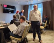 Eine Gruppe von Menschen nimmt an einem Geschäftstreffen in einem Konferenzraum der Hochschule Coburg teil. Einige sitzen und prüfen Dokumente, während eine Person in die Kamera blickt. Auf einer Leinwand ist eine Präsentationsfolie mit dem Titel „Veränderungen der Risikobereitschaft in den goldenen Jahren“ zu sehen.