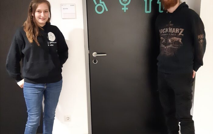 Zwei Personen stehen auf beiden Seiten einer Tür an der Hochschule Coburg, die mit Symbolen für inklusive Toiletten wie Rollstuhlfahrer, geschlechtsneutrale Toiletten und barrierefreie Toiletten geschmückt ist. Die linke Person trägt einen dunklen Kapuzenpullover und Jeans, während die rechte Person spiegelbildlich einen dunklen Kapuzenpullover und eine Hose trägt.