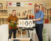In einer geschäftigen Werkstatt der Hochschule Coburg stehen zwei Männer zwischen Werkzeugen, die die Wände säumen. Der Mann links hält einen Blumenstrauß in der Hand, während sein Gegenüber neben ihm grinsend ein Schild mit der Aufschrift „1000“ hochhält. Sie lächeln strahlend neben einem Tisch, auf dem eine kleine Säge ausgestellt ist.