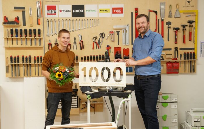In einer geschäftigen Werkstatt der Hochschule Coburg stehen zwei Männer zwischen Werkzeugen, die die Wände säumen. Der Mann links hält einen Blumenstrauß in der Hand, während sein Gegenüber neben ihm grinsend ein Schild mit der Aufschrift „1000“ hochhält. Sie lächeln strahlend neben einem Tisch, auf dem eine kleine Säge ausgestellt ist.