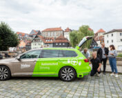 Ein grün-weißes Auto mit der Aufschrift „Valeo Automated Driving“ steht auf einer Kopfsteinpflasterstraße in der Nähe der Hochschule Coburg. Vier Personen stehen um den offenen Kofferraum herum und untersuchen die Ausrüstung. Im Hintergrund ist eine malerische Stadt mit Ziegeldächern und Bäumen zu sehen.