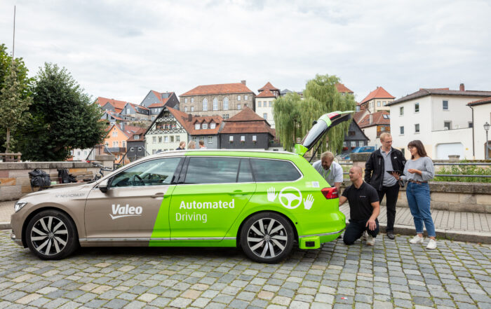 Ein grün-weißes Auto mit der Aufschrift „Valeo Automated Driving“ steht auf einer Kopfsteinpflasterstraße in der Nähe der Hochschule Coburg. Vier Personen stehen um den offenen Kofferraum herum und untersuchen die Ausrüstung. Im Hintergrund ist eine malerische Stadt mit Ziegeldächern und Bäumen zu sehen.