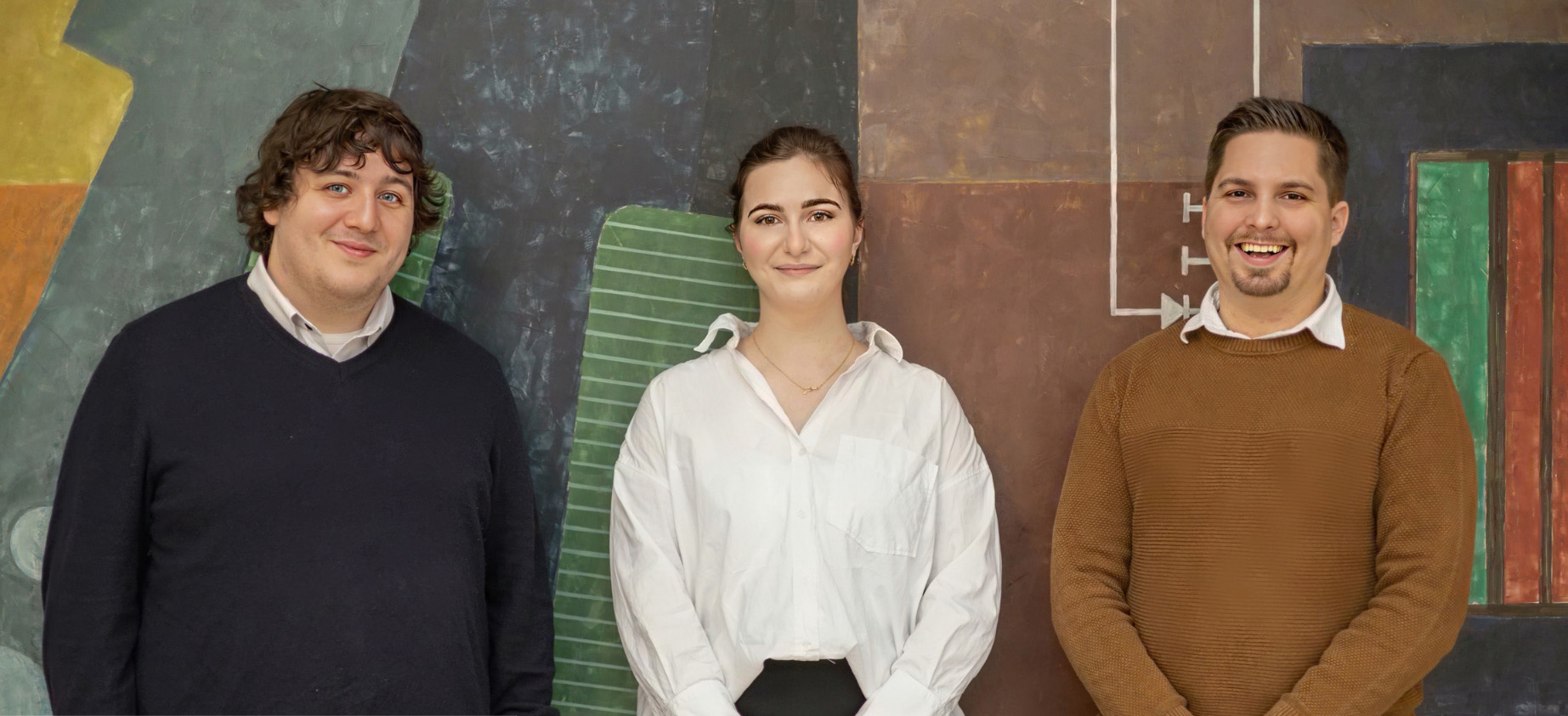 Vor einem farbenfrohen geometrischen Wandbild stehen stolz drei Personen von der Hochschule Coburg. Die Person links trägt einen dunklen Pullover, die Person in der Mitte ein frisches weißes Hemd und die Person rechts einen kuscheligen braunen Pullover. Alle lächeln strahlend in die Kamera.