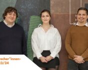 Drei Personen stehen lächelnd vor einem abstrakten Wandbild an der Hochschule Coburg. Die Person links trägt einen dunklen Pullover mit weißem Hemd, die Person in der Mitte eine weiße Bluse und einen schwarzen Rock und die Person rechts einen braunen Pullover mit Jeans. Deutscher Text ist sichtbar.