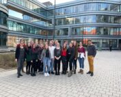 Auf einem gepflasterten Hof vor einem modernen Glasbau der Hochschule Coburg steht eine Gruppe von Menschen zusammen. Sie sind leger gekleidet, der Himmel ist klar.