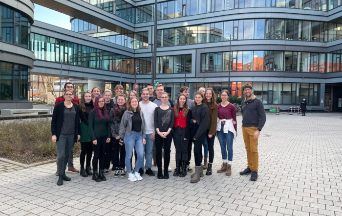 Auf einem gepflasterten Hof vor einem modernen Glasbau der Hochschule Coburg steht eine Gruppe von Menschen zusammen. Sie sind leger gekleidet, der Himmel ist klar.