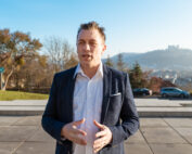 Ein Mann in weißem Hemd und dunkler Jacke steht draußen und gestikuliert mit den Händen. Hinter ihm ist der malerische Blick auf eine Stadtlandschaft mit Hügeln und einer Burg unter dem klaren blauen Himmel zu sehen, der an Aussichten in der Nähe der Hochschule Coburg erinnert.
