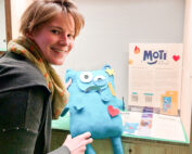 Eine Person lächelt und hält ein blaues Plüschtier mit Stern und Herz in der Hand vor einer Informationsvitrine der Hochschule Coburg.