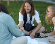 Drei Menschen sitzen auf der Wiese der Hochschule Coburg, unterhalten sich und schreiben auf Papier. Ein Mann mit langen Haaren lächelt, während zwei Frauen, eine davon mit Brille, aufmerksam zuhören. Sie befinden sich in einer entspannten Umgebung im Freien mit viel Grün im Hintergrund.