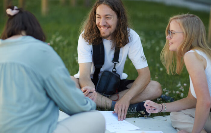 Drei Menschen sitzen auf der Wiese der Hochschule Coburg, unterhalten sich und schreiben auf Papier. Ein Mann mit langen Haaren lächelt, während zwei Frauen, eine davon mit Brille, aufmerksam zuhören. Sie befinden sich in einer entspannten Umgebung im Freien mit viel Grün im Hintergrund.