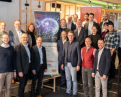 Eine Gruppe von Menschen, darunter Männer und Frauen, posiert lächelnd neben einem Banner zum Thema Künstliche Intelligenz an der Hochschule Coburg. Sie befinden sich in einer modernen Inneneinrichtung mit einer Treppe und einigen Plakaten im Hintergrund.