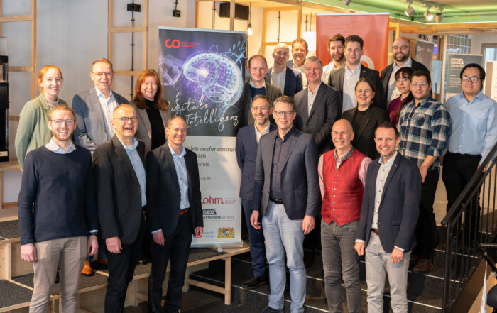 Eine Gruppe von Menschen, darunter Männer und Frauen, posiert lächelnd neben einem Banner zum Thema Künstliche Intelligenz an der Hochschule Coburg. Sie befinden sich in einer modernen Inneneinrichtung mit einer Treppe und einigen Plakaten im Hintergrund.