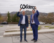Zwei Männer in Businesskleidung stehen draußen und halten ein Schild mit der Aufschrift „CO2 Science“. Sie lächeln und repräsentieren die Hochschule Coburg vor einer Kulisse aus Hügeln und Gebäuden. Der Himmel ist bedeckt, was der lebhaften Szene einen spannenden Kontrast verleiht.