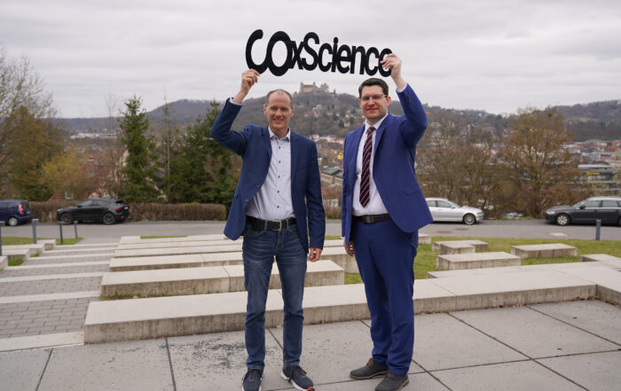 Zwei Männer in Businesskleidung stehen draußen und halten ein Schild mit der Aufschrift „CO2 Science“. Sie lächeln und repräsentieren die Hochschule Coburg vor einer Kulisse aus Hügeln und Gebäuden. Der Himmel ist bedeckt, was der lebhaften Szene einen spannenden Kontrast verleiht.