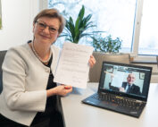Eine Frau lächelt, während sie ein unterschriebenes Dokument in der Hand hält, und ist sichtlich begeistert von ihrer Leistung. Sie sitzt an einem Tisch neben einem Laptop, auf dem ein Videoanruf mit einem Mann läuft. Beide zeigen stolz dasselbe Dokument der Hochschule Coburg. Eine Pflanze und ein Fenster bereichern die fröhliche Atmosphäre im Hintergrund.