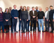 Eine Gruppe von sechzehn Personen steht in einem Flur mit rotem Boden und weißen Wänden der Hochschule Coburg. Sie sind leger gekleidet, lächeln und blicken in die Kamera.