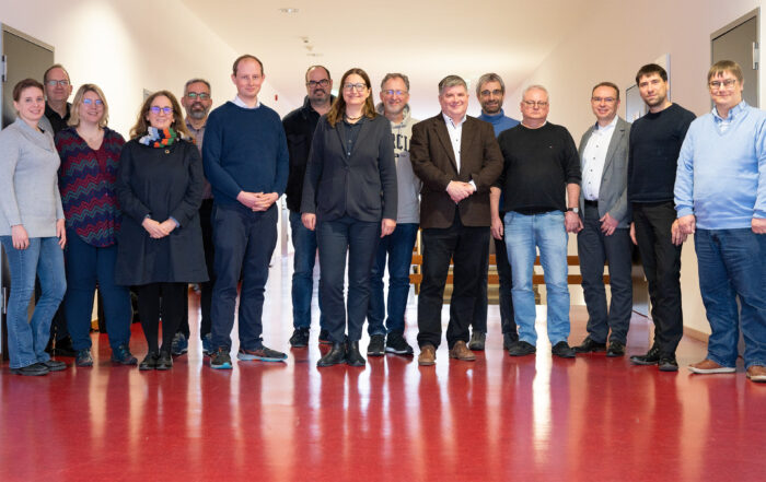 Eine Gruppe von sechzehn Personen steht in einem Flur mit rotem Boden und weißen Wänden der Hochschule Coburg. Sie sind leger gekleidet, lächeln und blicken in die Kamera.