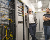 Zwei Männer stehen in einem Serverraum der Hochschule Coburg. Einer im Anzug lehnt an den Server-Racks, der andere steht im NASA-T-Shirt lässig mit verschränkten Armen inmitten eines Meeres aus Kabeln und Computerausrüstung.