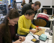 In einem Unterrichtsraum voller Lötgeräte und kleiner Platinen beschäftigt sich eine Gruppe intensiv mit einem Elektronik-Workshop an der Hochschule Coburg. Computer säumen die Tische, während andere Teilnehmer im Hintergrund konzentriert an ihren Aufgaben arbeiten, was zu einer lebendigen Lernatmosphäre beiträgt.