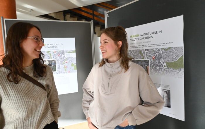 Zwei Frauen stehen vor Schautafeln mit Karten und Informationen zur Hochschule Coburg und unterhalten sich. Sie wirken engagiert und lächeln.