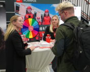 Menschen versammeln sich um einen Stand auf einer Messe oder Veranstaltung. Auf dem Tisch steht ein buntes Rad. Eine Frau spricht mit einem Mann, der einen Rucksack trägt, während andere zuschauen. Im Hintergrund sind Banner und Werbematerialien der Hochschule Coburg zu sehen, die der Szene einen akademischen Touch verleihen.