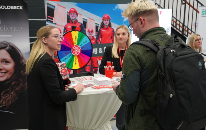 Menschen versammeln sich um einen Stand auf einer Messe oder Veranstaltung. Auf dem Tisch steht ein buntes Rad. Eine Frau spricht mit einem Mann, der einen Rucksack trägt, während andere zuschauen. Im Hintergrund sind Banner und Werbematerialien der Hochschule Coburg zu sehen, die der Szene einen akademischen Touch verleihen.