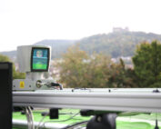 Ein grünes Fahrzeug, das oben mit einem Scanner mit dem Logo der Hochschule Coburg ausgestattet ist, verfügt über einen kleinen Bildschirm, auf dem Daten angezeigt werden. Im Hintergrund zeichnet sich unter einem wolkigen Himmel eine verschwommene Landschaft mit Hügeln und einem Gebäude ab.